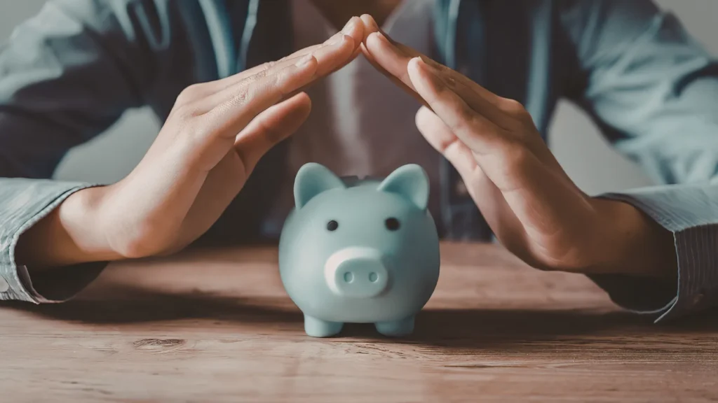 Primeiro plano de uma pessoa com as mãos em forma de telhado sobre um cofrinho azul em uma mesa de madeira, simbolizando proteção financeira e segurança das economias.